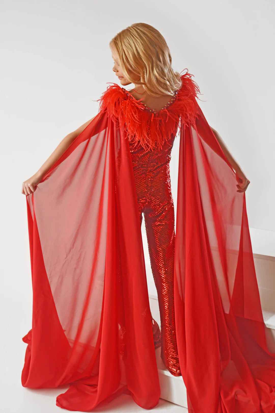 Ava Presley 38046 Long Sequin Jumpsuit is a delightfully fun fashion choice for girls. The detachable capes and feather bodice add playful panache for a statement-making look. The perfect pick for any occasion.  Sizes: 2-16  Colors: Red, Purple, White, Powder Blue